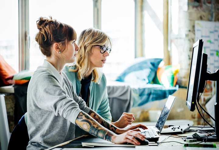 Two women collaborating on a computer in a modern office setting, focused on their work and sharing ideas.