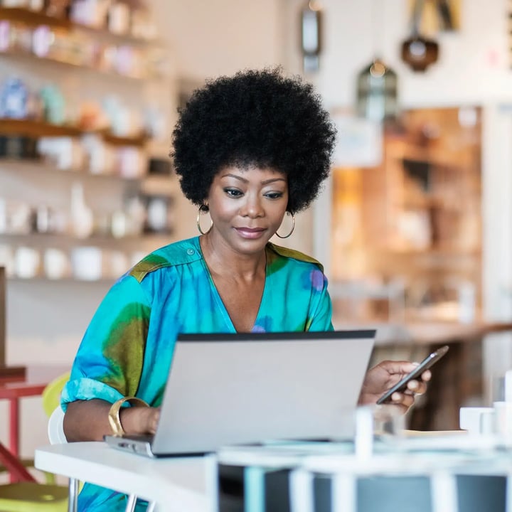 A woman with afro hair is focused on her laptop, engaged in work or study in a bright, modern environment.