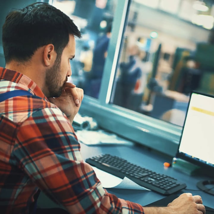 A man in a plaid shirt sits at a desk, focused on his computer screen, surrounded by a tidy workspace.