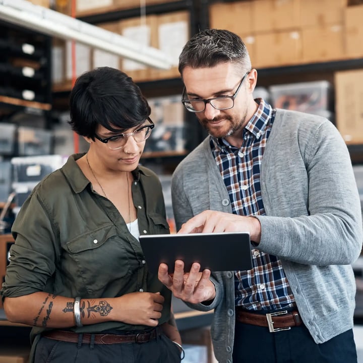A man and woman stand in a warehouse, examining a tablet together, surrounded by shelves and industrial equipment.