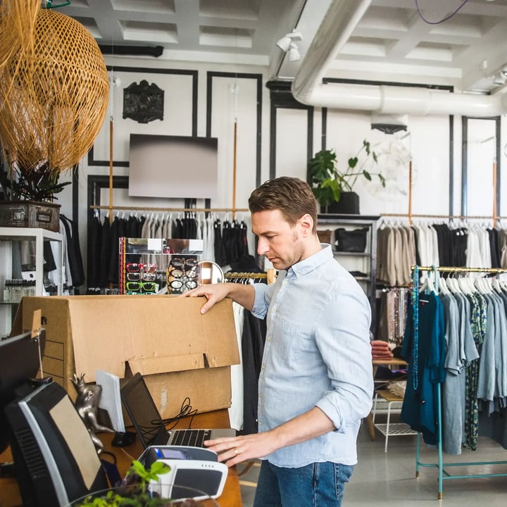 A man is focused on his laptop while seated in a clothing store, surrounded by various garments and displays. 
