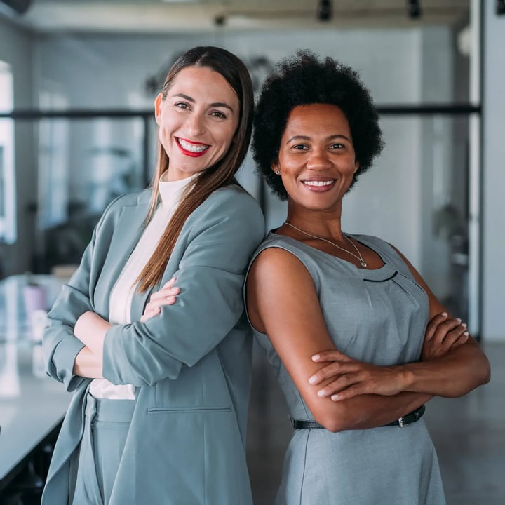 Two women smiling and posing back to back, both wearing gray outfits, in a modern office setting.