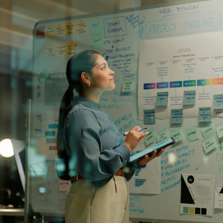 Women holds tablet and pen while staring up are white board with written ideas.