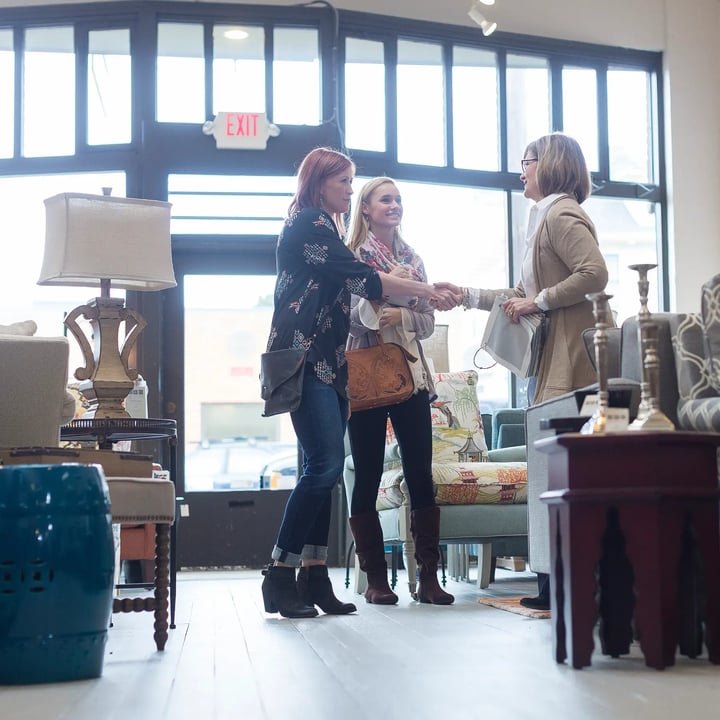 Three women stand together in a furnished room, engaged in conversation amidst the decor and furniture around them.
