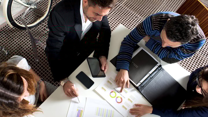 A diverse group of individuals gathered around a table, engaging with a laptop and a tablet in a collaborative setting.