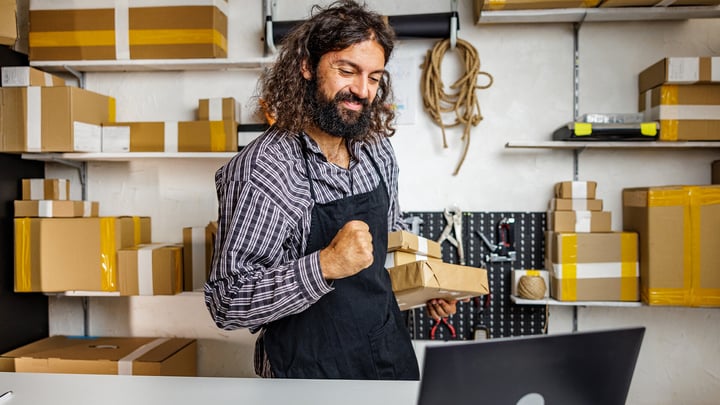 A smiling man with a beard, wearing an apron, holds a box in his hand, exuding warmth and friendliness.