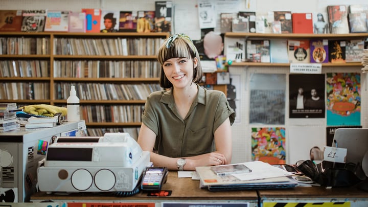 A woman seated at a desk in a record store, surrounded by vinyl records and music memorabilia, engaged in her work.