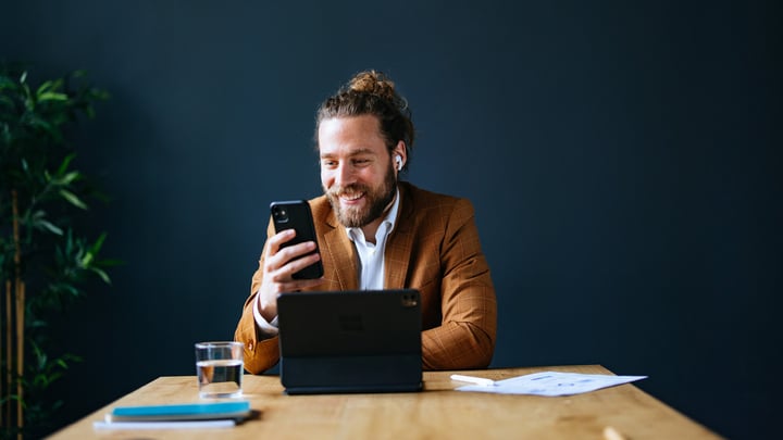 A man working in an office, using a laptop while simultaneously engaging with a smartphone.