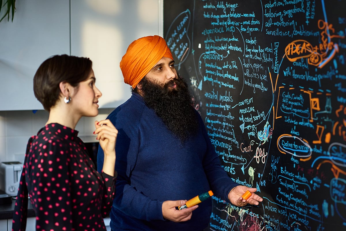 A man wearing an orange turban speaks with a female colleague in front of a black chalkboard. 