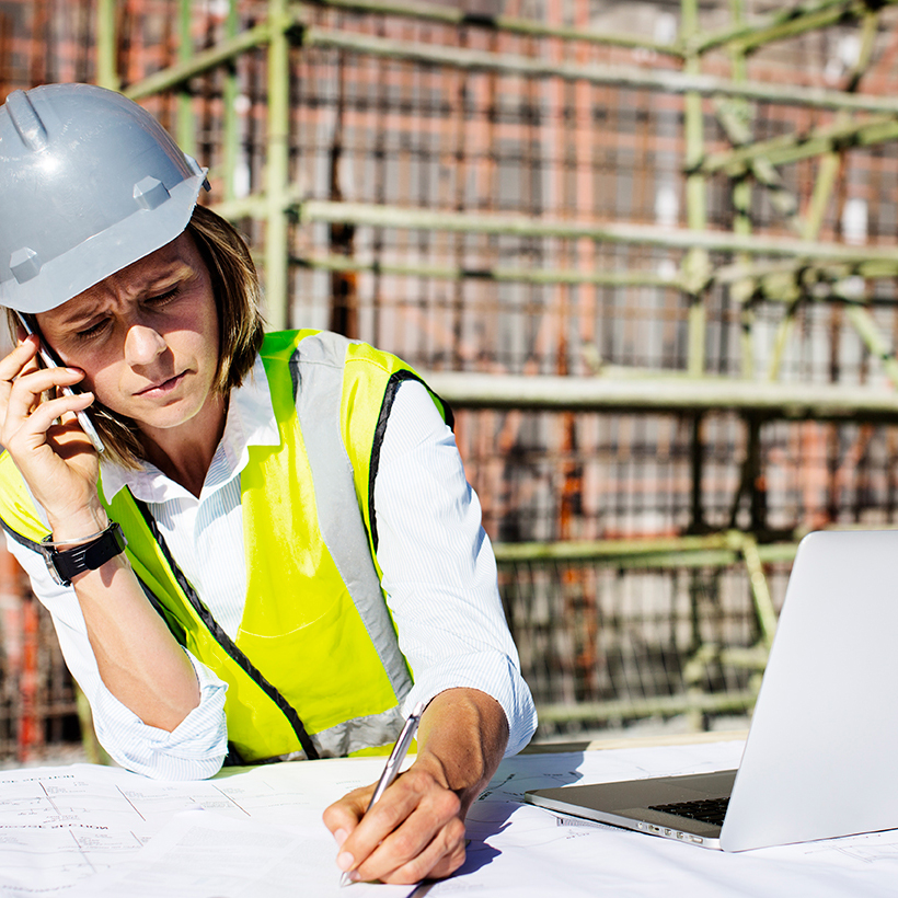 A female construction worker speaks on a phone about loan application while working on blueprints