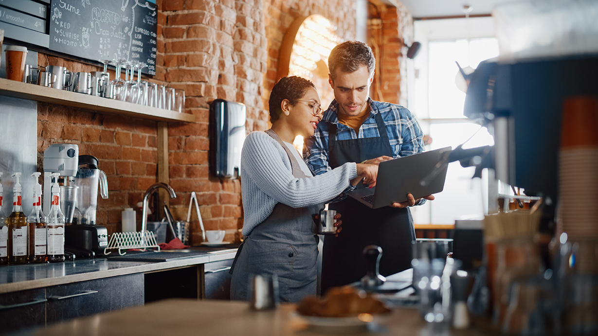 Two cafe employees wearing aprons looking at a laptop in a coffee shop.