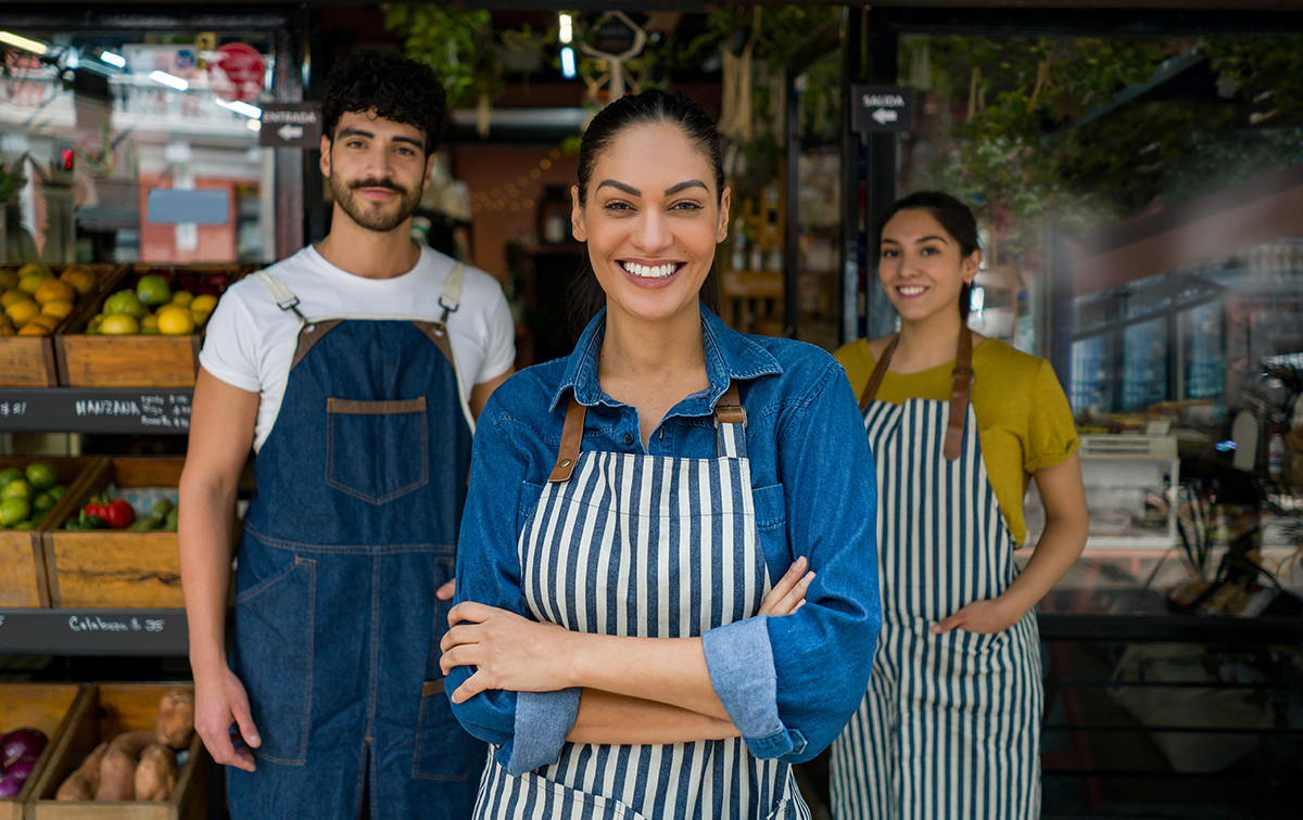 A manager wearing an apron stands in front of two employees wearing aprons in a grocery store. 